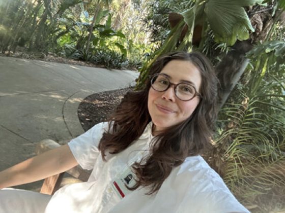 A selfie of a smiling woman with long brown hair and glasses sitting on an outdoor bench. She is wearing a crisp white short-sleeved shirt, and the background shows a dappled, sunlit path surrounded by dense, tropical garden foliage.