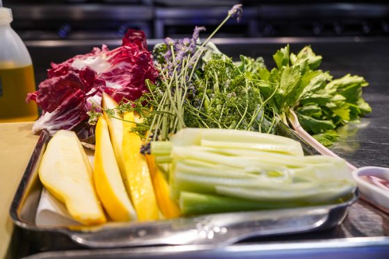 A stainless steel prep tray in a professional kitchen piled with fresh, vibrant ingredients ready for assembly, featuring thick wedges of yellow summer squash, stalks of celery, leafy green herbs, bitter radicchio, and delicate sprigs of purple lavender flowers.