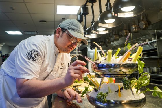 Chef George Murkowitcz, wearing a white chef's uniform, glasses, and a grey cap, meticulously arranges sprigs of lavender onto a multi-tiered silver display filled with ice and fresh raw vegetables. The shot is captured in a professional kitchen setting under bright overhead heat lamps.