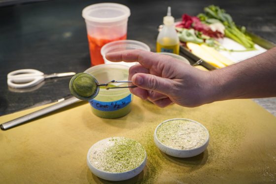 Close-up of a chef's hand dusting a fine green herb powder using a small mesh strainer over two shallow, round white bowls containing a creamy, light-colored base. In the background, prep containers, kitchen shears, a squeeze bottle, and an assortment of fresh raw vegetables are visible on a stainless steel workspace.