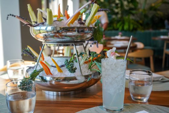 A two-tiered silver serving tower filled with shaved ice and an assortment of vibrant, freshly cut summer vegetables, including raw broccoli florets, carrots, and cucumber slices, interspersed with sprigs of lavender. The elegant presentation sits on a wooden dining table alongside a tall glass filled with crushed ice, a clear beverage, and a metal straw, with a blurred restaurant interior in the background.