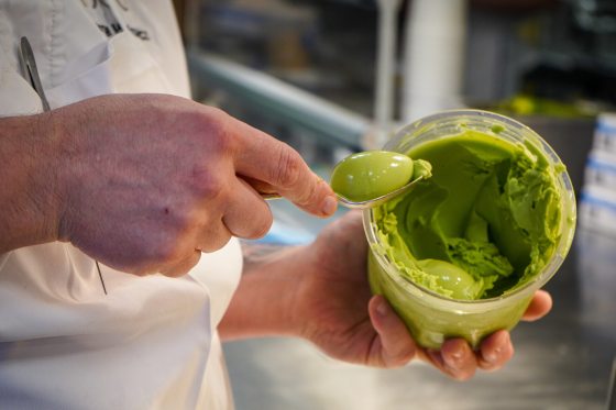 A close-up of a chef’s hands using a spoon to neatly scoop a smooth, vibrant green sorbet from a clear plastic storage container in a commercial kitchen.