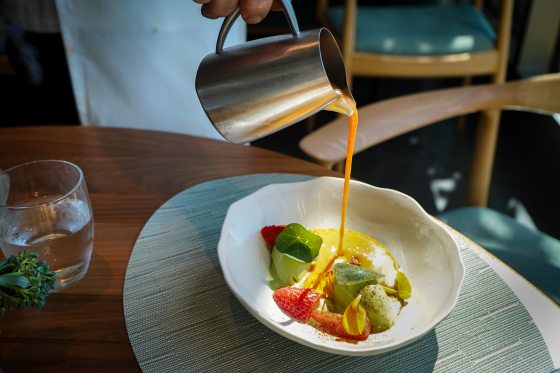 A close-up action shot of a bright orange gazpacho soup being poured from a small stainless steel pitcher into a shallow, wavy white bowl. The bowl is beautifully plated with scoops of green avocado sorbet or mousse, fresh sliced strawberries, a nasturtium leaf, and edible flower petals.