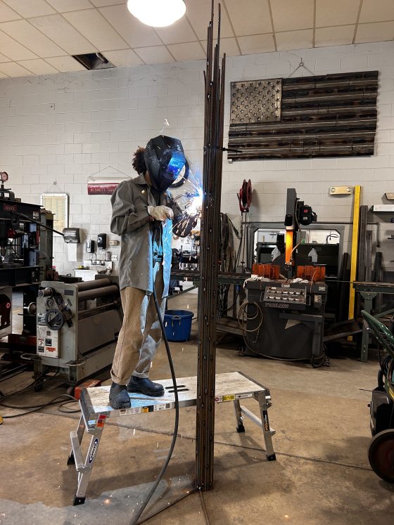 A person in a welding mask, welding a tall garden feature. 