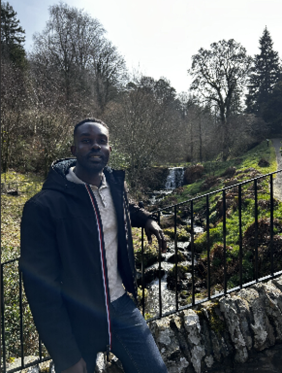 A person in a black jacket and jean, resting against a metal railing that overlooks a small waterfall.