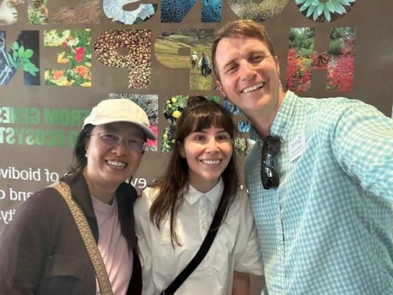A bright, smiling selfie of three people standing indoors in front of a graphic museum wall display. On the left, a woman wears a baseball cap and glasses; in the middle, a woman with bangs smiles warmly; on the right, a man in a blue gingham button-down shirt holds the camera.