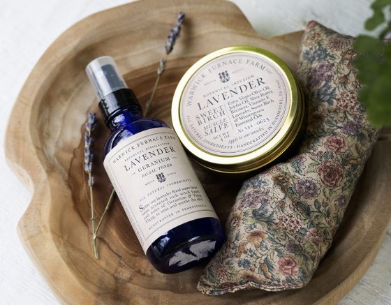 A flat-lay photograph on a white wooden surface featuring a wooden tray holding a blue glass bottle of Lavender Geranium facial toner, a tin of Lavender Sweet Birch muscle salve, a small floral-patterned sachet, and a wooden scrub brush.