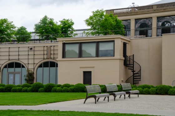 Exterior view of the Main Fountain Control Tower featuring a beige concrete building with a row of upper windows, a black spiral staircase, a neat green lawn, and several rows of pruned hedges under an overcast sky.