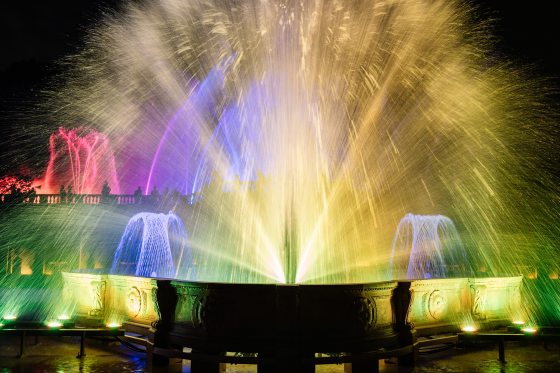 A vibrant night view of an illuminated fountain show featuring powerful central jets lit in bright yellow and surrounded by streams of blue, purple, and green light against a dark background.