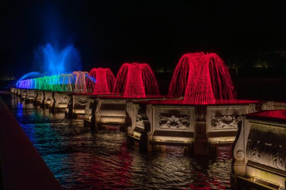 A perspective view along a pool of water at night, showcasing a sequence of ornate stone fountain basins illuminated in alternating bold red, green, and blue colors with bright blue streams shooting high in the background.