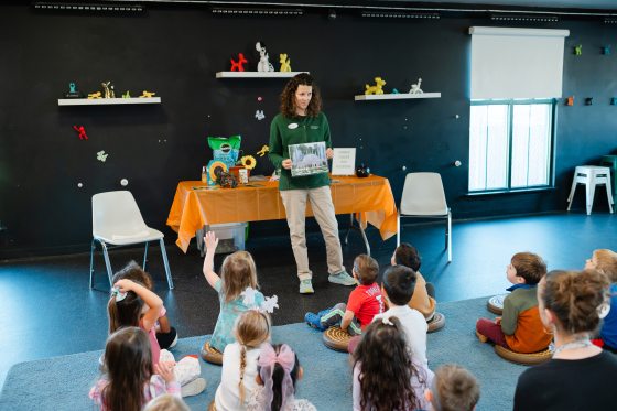 An adult educator stands in a classroom, holding a picture of a garden conservatory, as children sit on the floor listening and participating in a lesson.