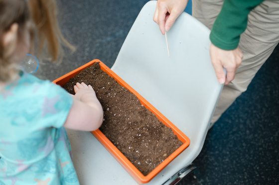 A child’s hand touching the soil in a rectangular orange planter, with an adult instructor nearby holding a small wooden dowel.
