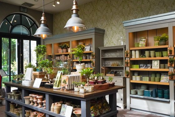A well-lit garden shop interior featuring a central wooden table display of various bonsai trees and gardening books, with shelving units in the background stocked with ceramic pots, plants, and additional retail items under industrial-style pendant lighting.