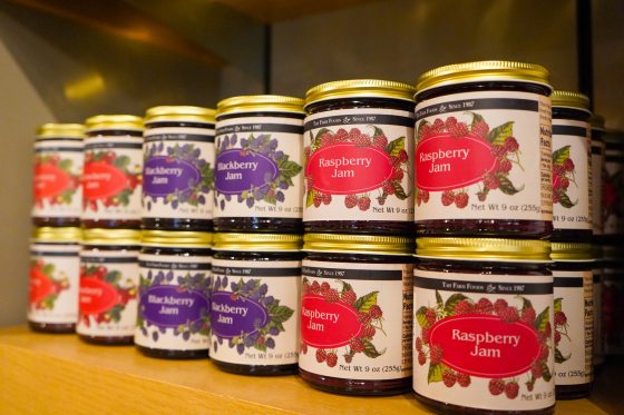Rows of jam jars arranged on a wooden shelf, featuring labels for Raspberry Jam and Blackberry Jam with illustrative berry graphics on the front.