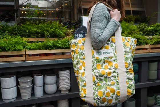 A person carrying a large canvas tote bag featuring a green and yellow lemon and foliage pattern, with the Longwood Gardens logo clearly visible on the front patch, while standing in a garden shop filled with plants and pottery.