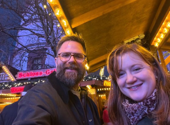 A selfie of a smiling couple standing outdoors at night under string lights in front of a German-style market stall with a sign that reads "Schloss-Grill".