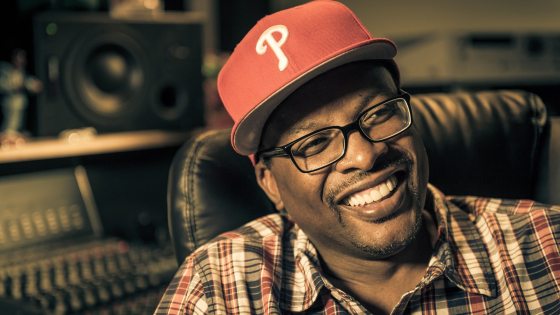 A close-up portrait of DJ Jazzy Jeff smiling broadly, wearing glasses, a plaid shirt, and a red baseball cap with a white "P" logo inside a recording studio.