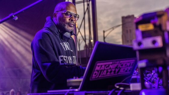 DJ Jazzy Jeff standing behind a DJ setup outdoors at dusk, wearing a dark hoodie and glasses, with a laptop sticker reading "The Magnificent DJ Jazzy Jeff."