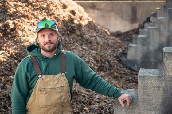 A man with a beard and baseball cap wearing a green hoodie and tan overalls stands outdoors near a large pile of wood chips and concrete structures.