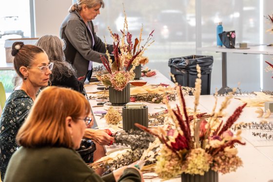 Several participants are seated at long tables working on dried floral arrangements containing textured burgundy stalks, grasses, and hydrangeas in dark ribbed containers during a continuing education class.