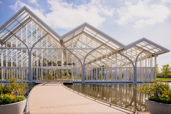 The modern glass and steel structure of the West Conservatory under a partly cloudy sky, featuring a curved concrete walkway crossing a still reflecting pool.