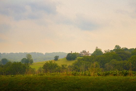 A wide landscape view of rolling green hills and dense forests under an expansive, hazy sky, with a large estate house nestled in the distance.