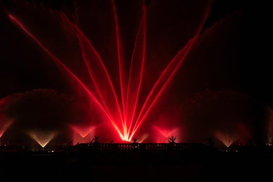 A nighttime outdoor fountain show features dramatic beams of bright red water shooting upward into the dark sky in a fan-like pattern from a stone terrace.
