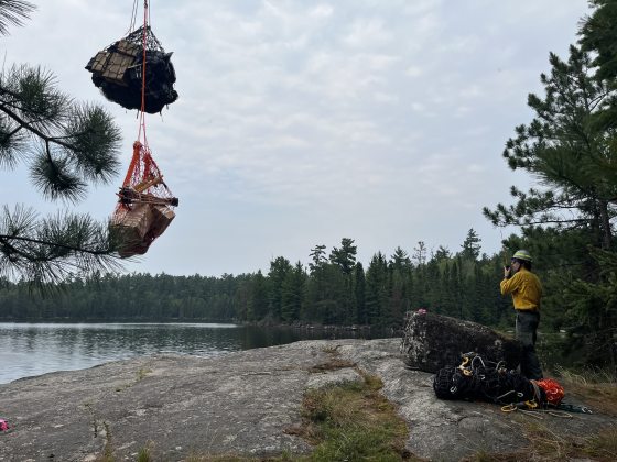 A firefighter stands on a rocky shoreline speaking into a radio, watching two heavy-duty cargo nets carrying boxed supplies dangle from a line attached to an aircraft overhead.