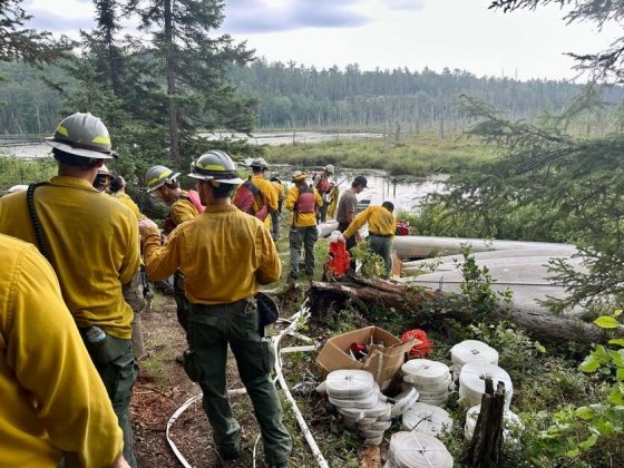 A group of firefighters in yellow jackets and hard hats stand near stacked canoes and rolls of white fire hoses on a grassy bank by a body of water.