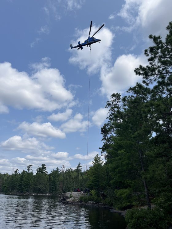 A blue helicopter flies against a partly cloudy sky, with a long line suspended from its underside extending down toward a tree line near a body of water.
