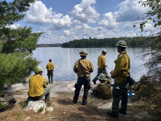 Several firefighters in yellow jackets and hard hats gather along a rocky shoreline, looking out over a calm lake surrounded by forested shores under a cloudy blue sky.