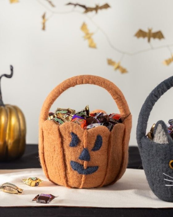 A felt pumpkin trick-or-treat basket filled with foil-wrapped candies sits on a table with scattered sweets, while a black cat basket and gold bat garlands decorate the white wall behind it.