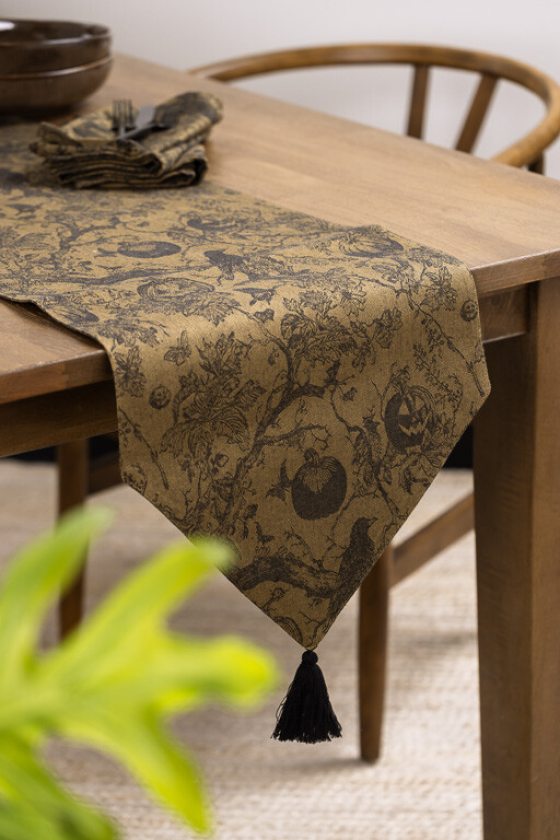 A wooden dining table is set with a dark, illustrated Halloween-themed table runner featuring crows, pumpkins, and bare trees, finished with a black tassel hanging off the edge.