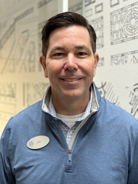 A man with black hair wearing a light blue shirt and a nametag smiles in front of a white wall with grey sketches on it. 