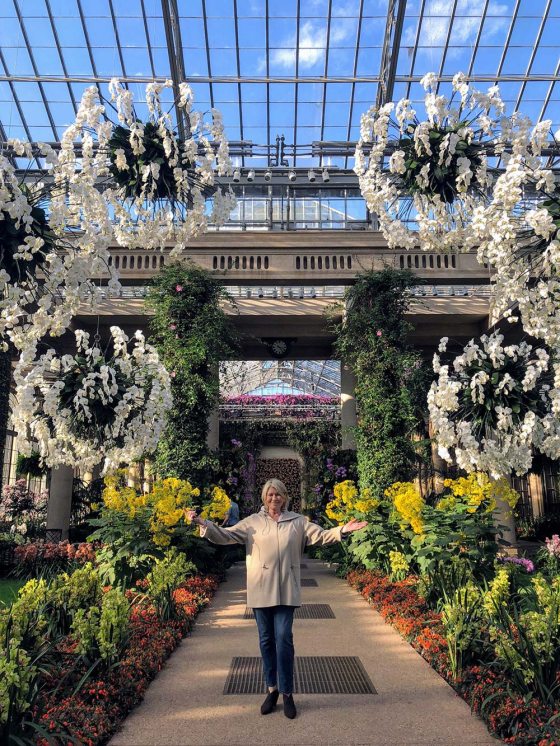 A person smiles with arms outspread  on a central walkway surrounded by conservatory plants and a glass roof