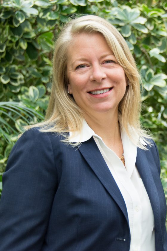 A person in a blue jacket and white shirt, with medium length blond hair, smiling at the camera.
