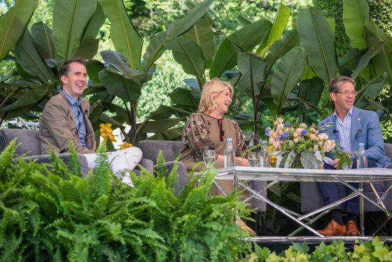 a group of three people sitting on an outdoor couch surrounded by plants 